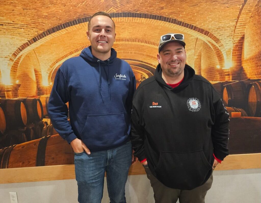 Paul Sharpe of Surfside Garage Doors and Dan Latini of Dan Can Home Services stand together smiling in front of a warm, barrel-vault brick backdrop during a local Ocean County community event.